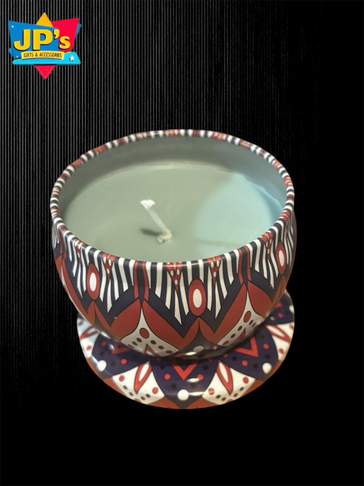 A candle in a patterned tin, set against a black textured background. The candle wax is a pale sage green, and the cup features a geometric pattern in red, white, and black.