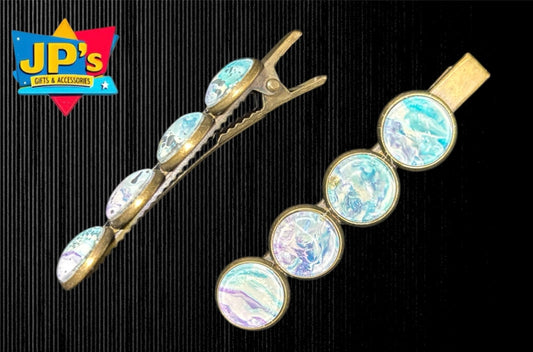 Two gold hair clips with blue and purple enamel patterns. One is a snap clip, the other is a round barrel clip style.