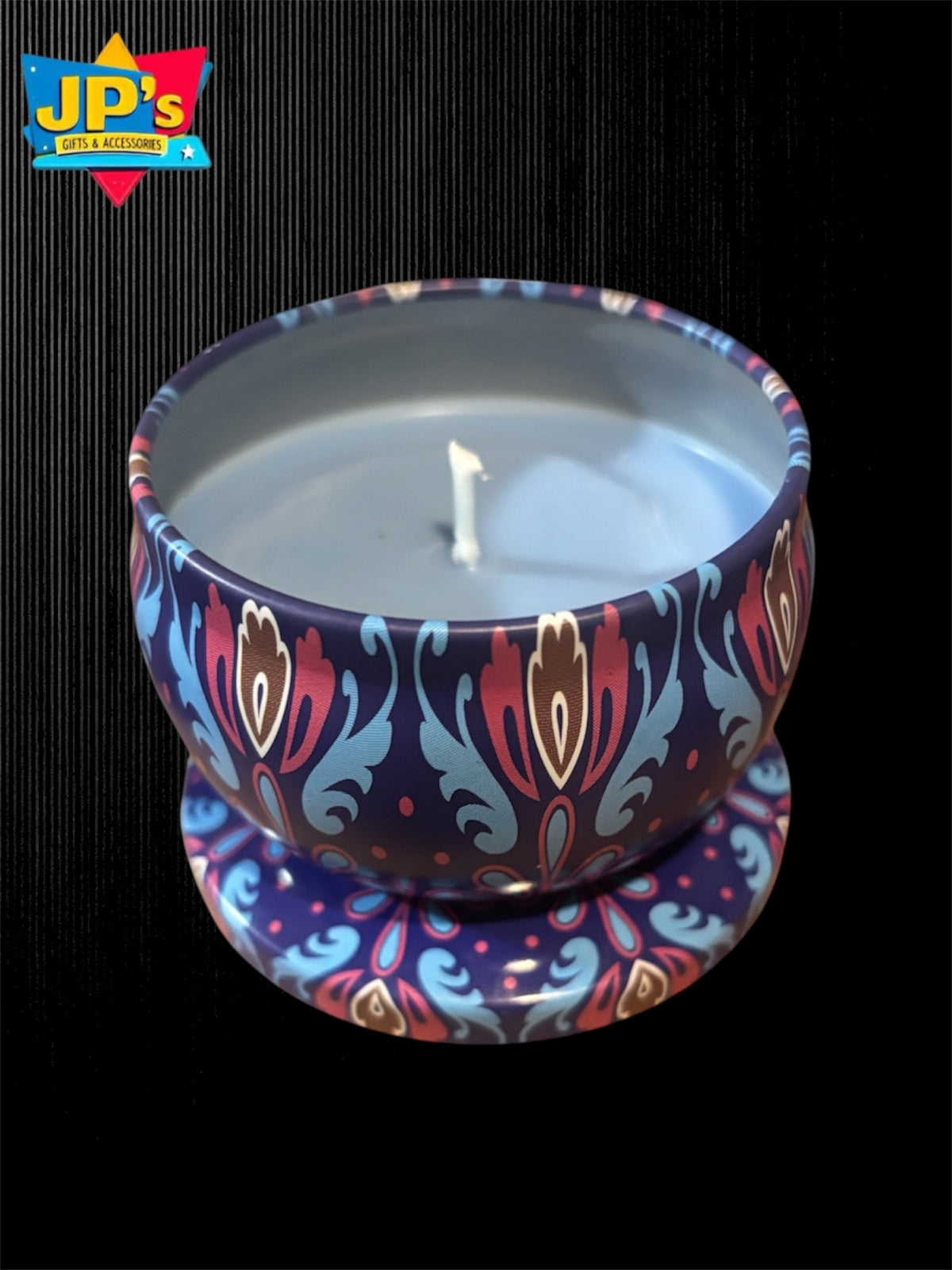 A two-piece candle set consisting of a round tin candle with a white wick and matching saucer, both decorated with a colorful paisley pattern in red, blue, and gold on a dark navy base. The candle is set against a black background with a JP's gifts and accessories logo in the top left corner.